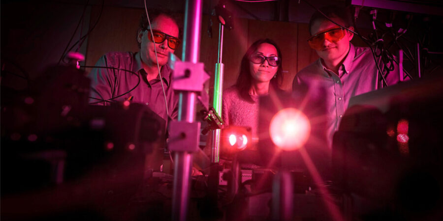 Three professionals operate a laser. The professionals and the room is bathed in a pink light.