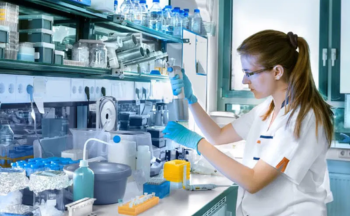 A woman sits in a lab and examines a beaker.