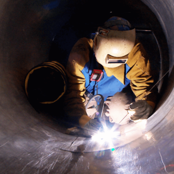A worker welds a pipe while inside an enclosed space.