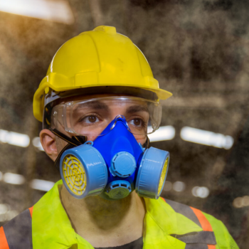 Close up an a worker wearing a respiratory mask, eyewear, and a hard hat. Dust fills the air.